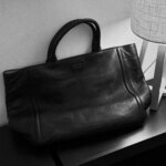 Black leather tote bag resting on a dark table beside a fabric-shade lamp, evoking a return to work and readiness for growth.