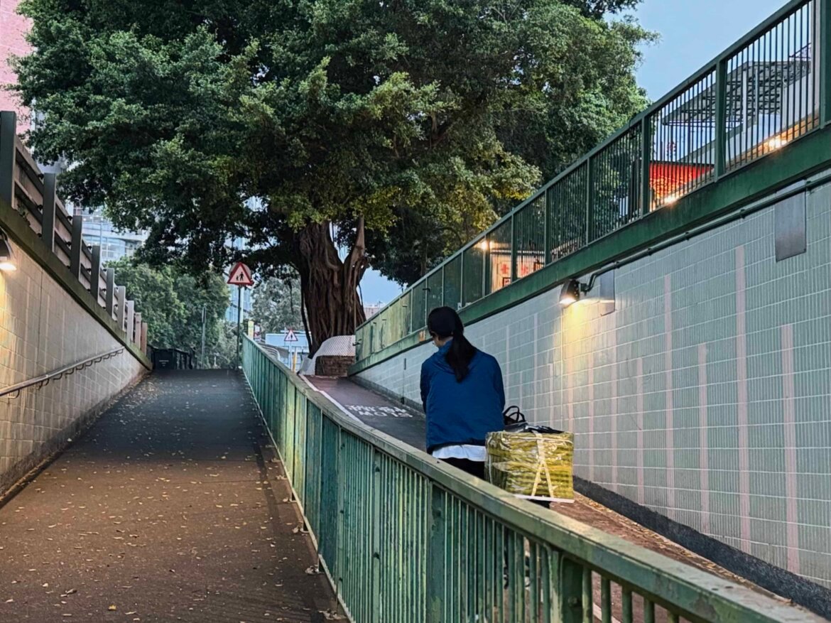 A woman seen from behind walking along a pedestrian ramp in Hong Kong at dusk, carrying a yellow bag, with a large banyan tree and green railings framing the path ahead.