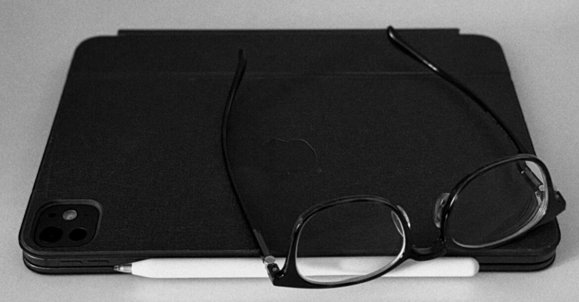 A black-and-white close-up of a tablet and a smartphone lying flat on a light surface, with a white stylus tucked along the bottom edge and a pair of black-framed eyeglasses resting on top. The tools of a one-person team, photographed in grayscale.