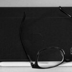 A black-and-white close-up of a tablet and a smartphone lying flat on a light surface, with a white stylus tucked along the bottom edge and a pair of black-framed eyeglasses resting on top. The tools of a one-person team, photographed in grayscale.