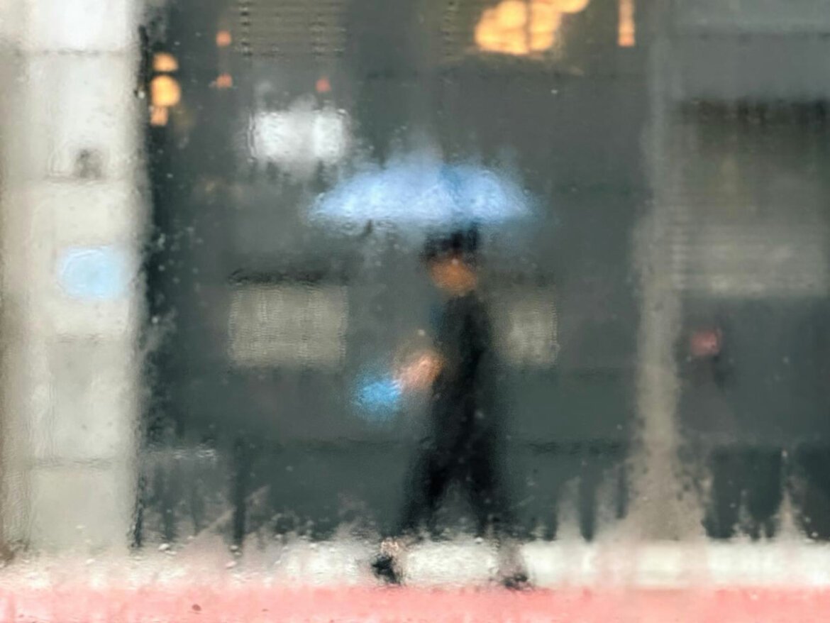 A rainy day in Hong Kong. A person walks along a wet sidewalk holding a light blue umbrella, bright blue bag in hand, warm shop lights glowing in the background. Saul Leiter style.