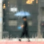 A rainy day in Hong Kong. A person walks along a wet sidewalk holding a light blue umbrella, bright blue bag in hand, warm shop lights glowing in the background. Saul Leiter style.